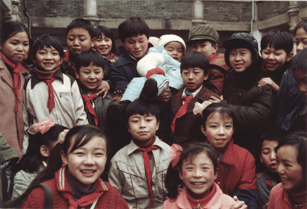 一群戴着红领巾的孩子们拥挤在一起合影，中间有一个被抱起的幼儿，背景是老式青砖建筑